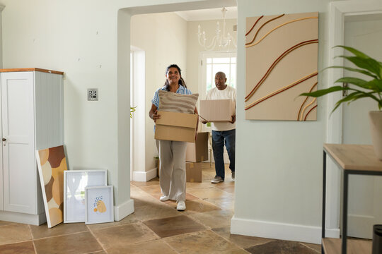 African American couple carrying moving boxes and striped cushion while entering tiled foyer