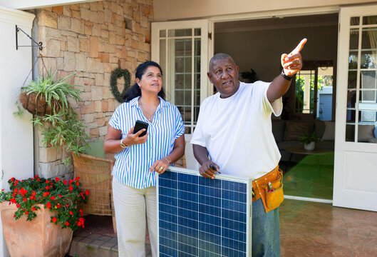 Mature woman and mature man standing on front porch with solar panel, tool belt, smartphone