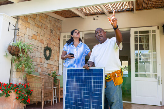 Mature man, woman holding solar panel, man wearing yellow tool belt, using phone on covered patio