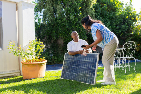 African American couple inspecting large solar panel and comparing smartphone data on backyard lawn
