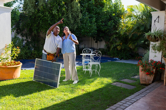 African American technician homeowner with toolbelt holding solar panel pointing upward in backyard