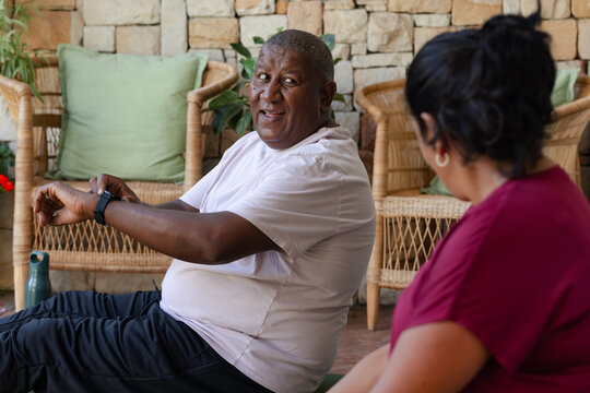 Senior African American couple sitting on yoga mats on covered patio, man checking smartwatch