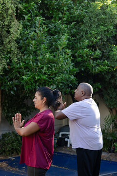 African descent senior couple standing praying in garden in maroon top, light tee near pool cover