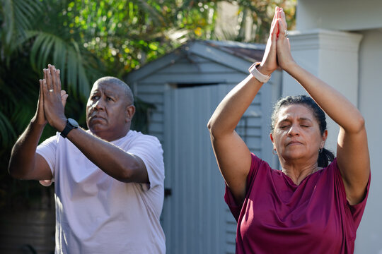 African American senior couple meditating in garden by gray shed, in short sleeves with watches