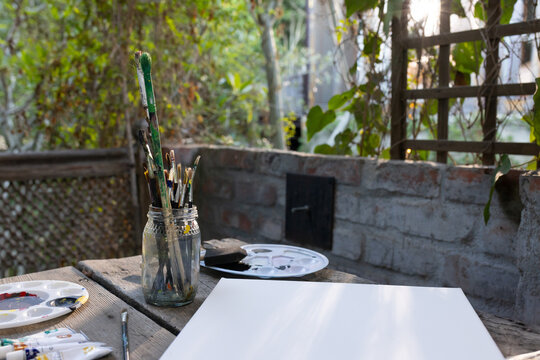 Glass jar holding paintbrushes is resting on weathered table in garden nook, sunlight dappled