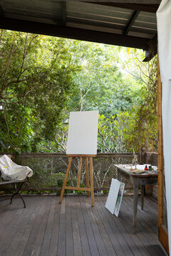 Wooden easel is holding blank white canvas on covered porch facing lush garden, with paint table