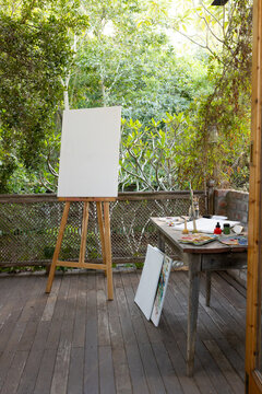 Wooden easel is holding blank stretched canvas on deck while paint palettes and brushes are waiting