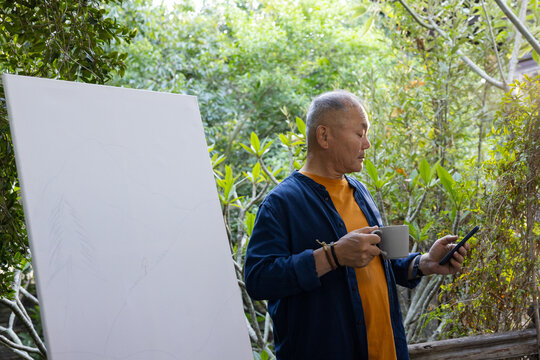 Senior Asian man standing on deck consulting smartphone and holding ceramic mug near blank canvas