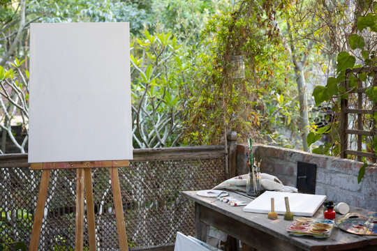 Primed canvas standing on easel on garden patio beside work table with brushes paints palette vines
