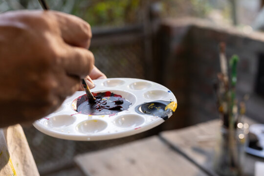 Mature adult painter hands mixing dark red and yellow paint with brush on white palette outdoors