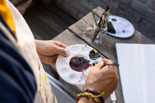 Senior Asian male wearing stained apron holding white palette mixing paint with thin brush at bench