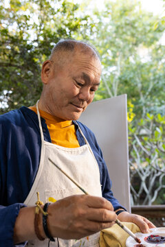 Senior Asian man painting at easel in garden, wearing mustard shirt and holding palette