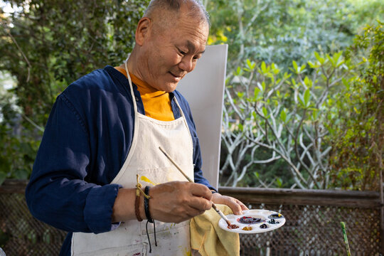 Senior Asian man painting at garden railing wearing white apron mixing paint on palette