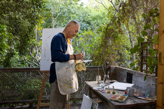 Senior Asian man standing at easel on garden porch, wearing paint-stained apron holding palette