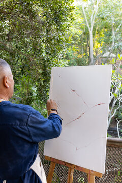 Senior Asian man wearing apron painting at easel on patio using brush on canvas, copy space