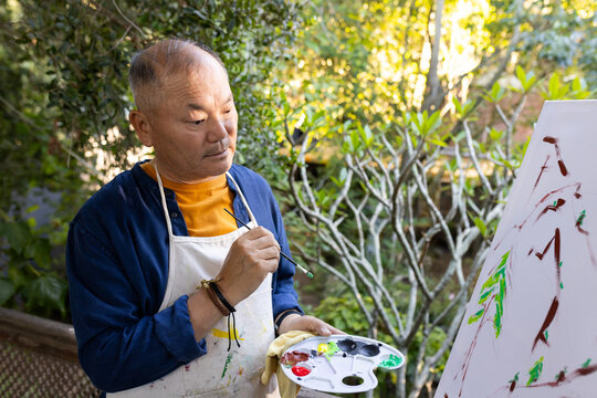 Senior Asian man wearing paint-stained apron holding palette and brush painting on canvas at patio