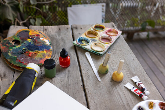 Two paint-smeared palettes and mixing tray are sitting on weathered wooden table on deck