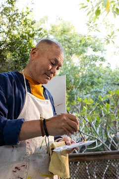 Senior Asian man standing on wooden deck mixing paint on palette while holding brush wearing apron