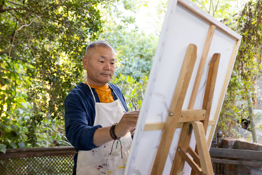 Senior Asian man in paint-stained apron standing on porch painting canvas on easel holding brush