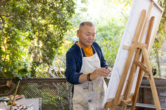 Senior Asian man painting canvas at easel on yard patio, wearing painted apron, holding palette