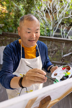 Senior Asian man painting on wooden deck at easel, holding palette and wearing paint-stained apron