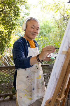 Senior Asian male wearing headset and paint-stained apron painting on deck at easel with palette