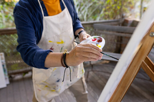 Senior Asian man painting on porch at easel wearing paint-stained apron holding palette