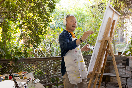 Senior Asian man standing at easel on porch painting canvas with palette, wearing headset and apron