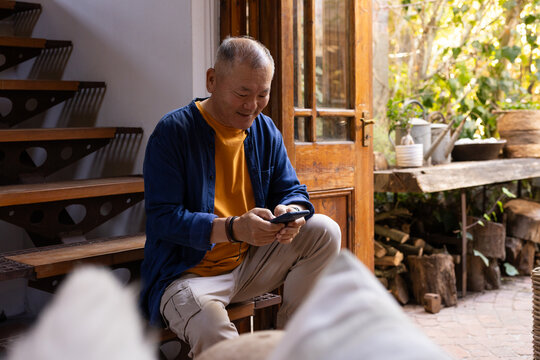 Senior Asian man sitting on low wooden step at home holding smartphone near open wooden door