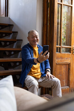 Senior Asian man holding smartphone while sitting on step by wood glass door near open stairs