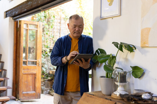 Senior Asian man in mustard tee and blue overshirt, standing at home holding tablet by Monstera
