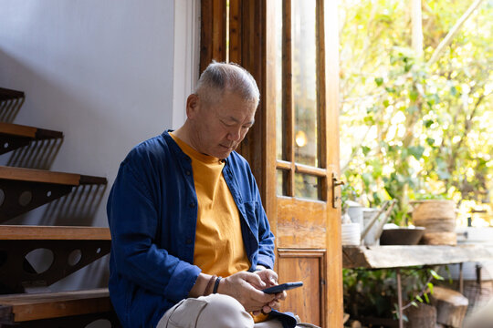 Senior Asian man sitting on wooden steps by open door, using smartphone, wearing mustard shirt