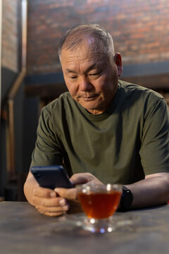 Senior Asian man sitting at wooden table in cafe in olive T-shirt holding smartphone with teacup