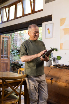 Senior Asian man standing in sunlit dining area beside wooden table holding tablet, tapping screen