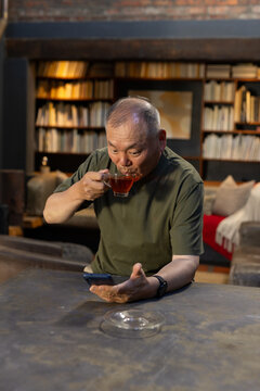 Senior Asian man olive tee at worn table home library, sipping glass teacup and using smartphone