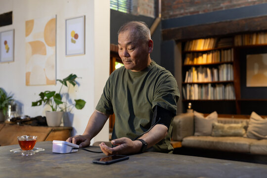 Senior Asian male checking blood-pressure using electronic monitor at wooden table in living room