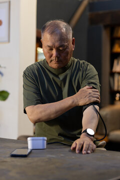 Senior Asian man sitting at home adjusting blood pressure cuff with white monitor, in olive shirt
