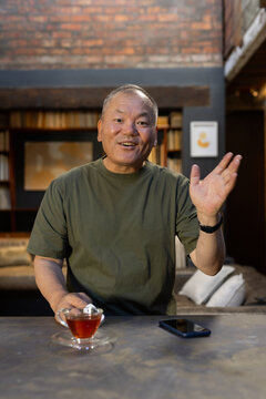 Senior Asian man waving and sitting at counter in study wearing olive tee with teacup smartphone