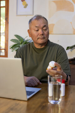 Senior Asian man wearing olive tee and watch examining pill bottle beside laptop at home table