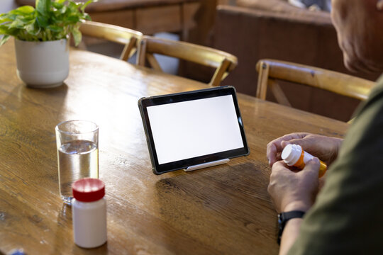 Senior man sitting at dining table holding pill bottle, reading label beside tablet, copy space