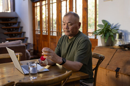 Senior Asian man handling pill container while sitting at dining table with laptop and water