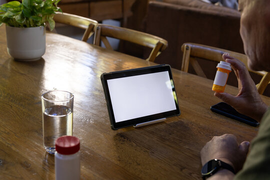 Senior Asian man sitting at wooden table holding amber pill bottle inspecting tablet, copy space