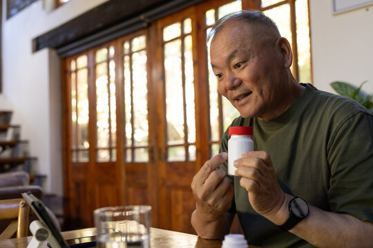 Senior Asian man holding and inspecting pill bottle with red cap at wooden table with tablet