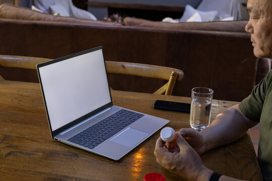 Senior Asian man in dark green tee holding orange pill bottle viewing blank laptop at table