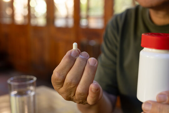 Senior Asian man holding white capsule near camera at kitchen table with pill bottle, glass