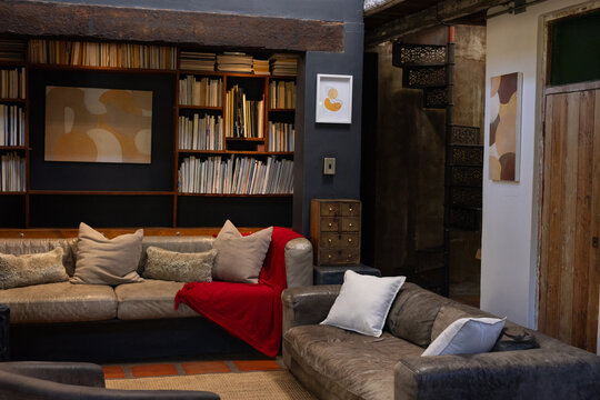 Two leather sofas are forming L-shape in rustic loft, featuring red throw, bookshelf, spiral stairs