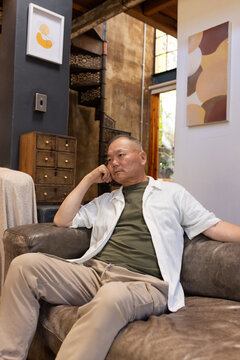 Senior Asian man sitting on brown leather sofa by metal spiral staircase, wearing olive green shirt