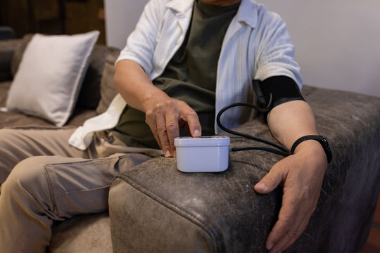 Senior man is measuring blood pressure on gray sofa in living room, with white monitor