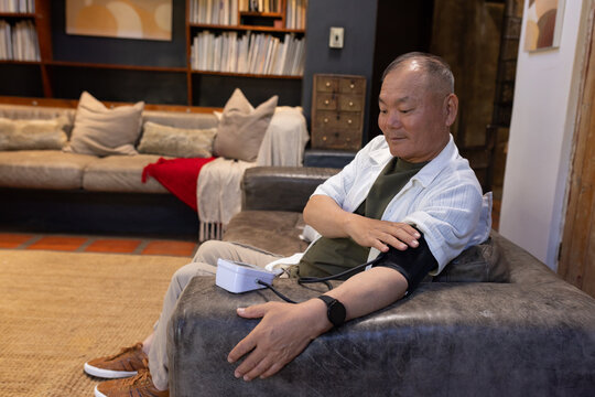 Senior Asian man sitting on dark gray armchair at home monitoring blood pressure cuff, copy space