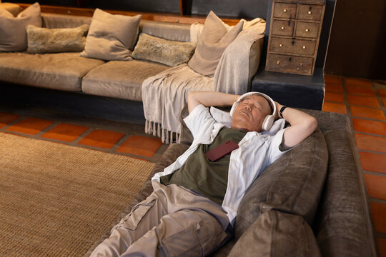Senior Asian man lying on chaise in lounge wearing headphones, phone resting on chest, copy space